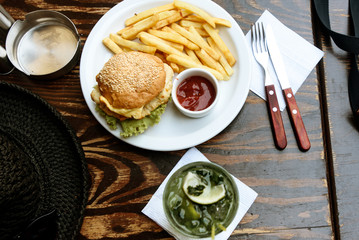delicious chicken burger with potato fries and ketchup and mojito cocktail on wooden table in restaurant
