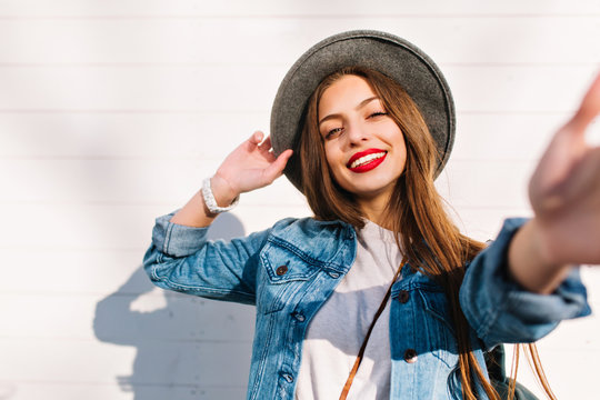 Cheerful brunette girl in white wristwatch and gray hat posing in front of wooden wall touching camera. Stylish smiling young woman with long brown hair and red lips having fun outside in sunny day