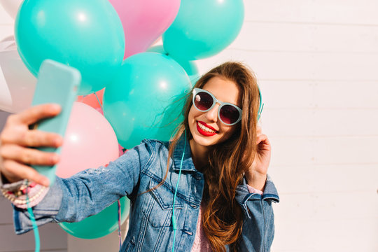 Adorable young woman with long hair makes new photo for facebook profile while having fun at friend's party. Gorgeous brunette girl in sunglasses and denim jacket takes selfie and enjoys festive event