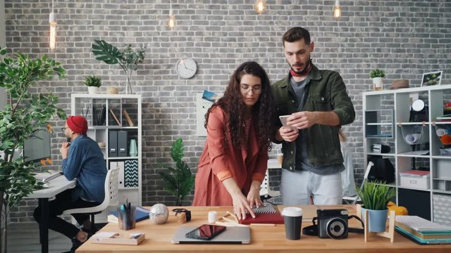 Girl is putting things on table making beautiful design while guy is shooting flat lay with smartphone camera holding device touching screen working in office together.