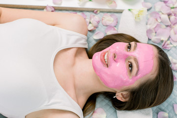 Obraz na płótnie Canvas Beautiful young woman is getting pink facial clay mask at spa, lying on bed with flower petals and fresh fruits