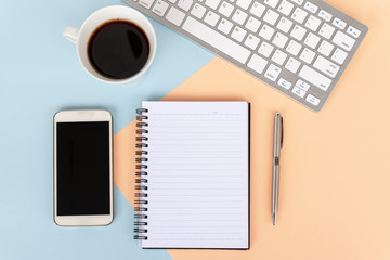 Directly above view of office desk with computer keyboard, cup of coffee,smart phone, note pad and pen on dual tones background