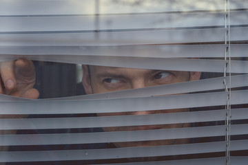 Man watching through window blinds Portrait of young thoughtful male with brown eyes observing through window jalousie