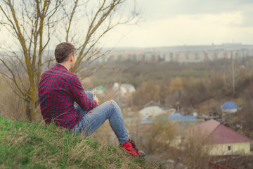 Naklejka na ściany i meble Young man sitting on green hill Side view of young male in jeans and plaid shirt sitting on grass and looking at camera