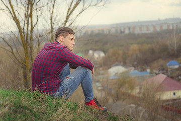 Young man sitting on green hill Side view of young male in jeans and plaid shirt sitting on grass and looking at camera