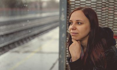 Dreamy young woman sitting beside railway Side view of young female in leather jacket sitting on background of railroad in overcast day