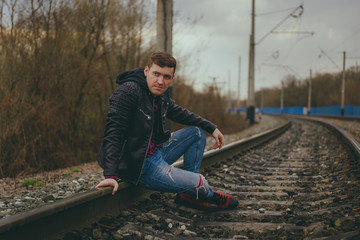 Trendy young man sitting on rails Pensive male in jeans and leather jacket sitting on railway in evening of cloudy day