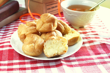 Bitten homemade small cakes profiterole choux pastry with custard next to a white cup with tea and books on a table with a rustic tablecloth in a red-white cage
