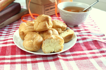 Bitten homemade small cakes profiterole choux pastry with custard next to a white cup with tea and books on a table with a rustic tablecloth in a red-white cage