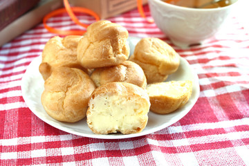 Bitten homemade small cakes profiterole choux pastry with custard next to a white cup with tea and books on a table with a rustic tablecloth in a red-white cage