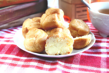 Bitten homemade small cakes profiterole choux pastry with custard next to a white cup with tea and books on a table with a rustic tablecloth in a red-white cage