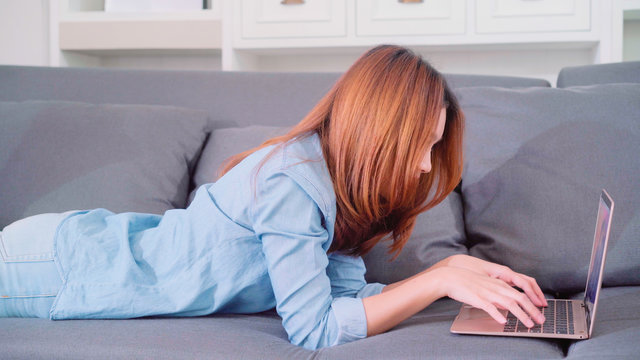 Portrait of beautiful attractive young smiling Asian woman using computer or laptop while lying on the sofa when relax in living room at home. Enjoying time lifestyle women at home concept.