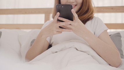 Young Asian woman using smartphone while lying on bed after wake up in the morning, Beautiful attractive Japanese girl smiling relax in bedroom at home. Enjoying time lifestyle women at home concept.