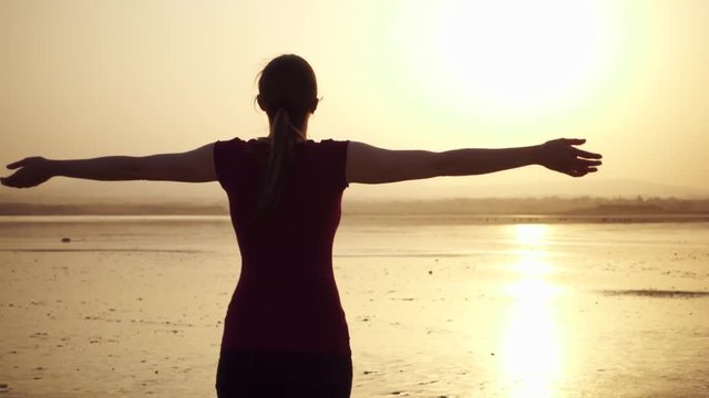 Dark silhouette of young woman raising arms up at sunset on lake. Behind view of female figure outstretching hands at golden hour in slow motion. Concept of new experience and freedom in traveling