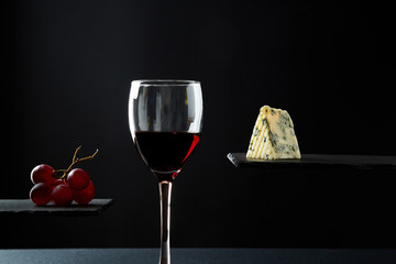 Composition of red wine in glass beside blue cheese and grape on black background.