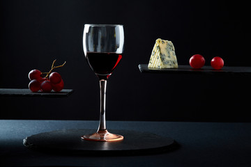 Composition of red wine in glass beside blue cheese and grape on black background.