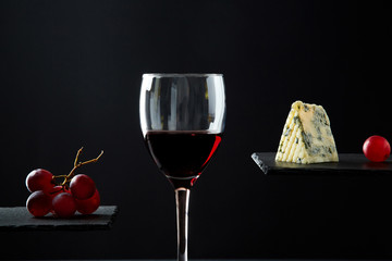 Composition of red wine in glass beside blue cheese and grape on black background.