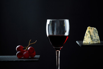 Composition of red wine in glass beside blue cheese and grape on black background.