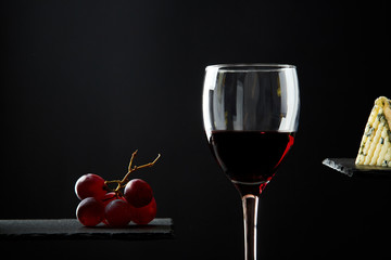 Composition of red wine in glass beside blue cheese and grape on black background.