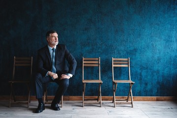 Waiting for interview. Confident mature businessman sitting on chair against dark blue background.