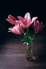 bouquet of pink lily flowers in the rays of light on a black background on a wooden rustic table in a glass bottle. fresh buds of a flowering plant close-up in a vase, copy space. studio shot