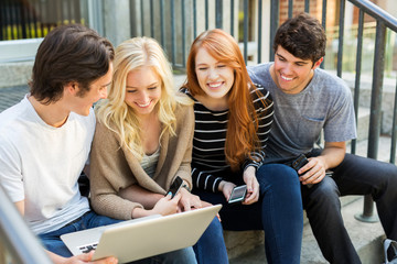 Four students sitting in a row on a step using their technology on the university campus and laughing together; Edmonton, Alberta, Canada