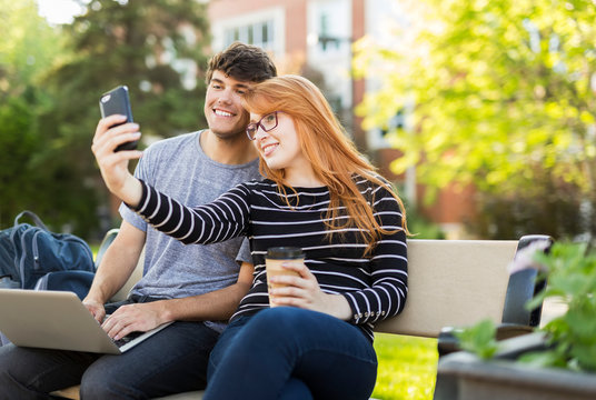 A young man and young woman sit together on a bench on the university campus taking a self-portrait on a smart phone while drinking coffee; Edmonton, Alberta, Canada