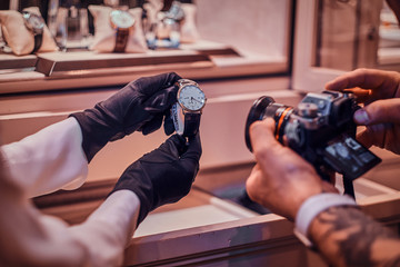 Tattoed photographer is taking photo of expensive watch in posh shop, while shop assistant is...
