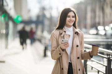 Young businesswoman checking email via mobile phone and holding a coffee cup against urban scene.