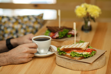 man takes a cup of coffee from a wooden table, on which lies a sandwich