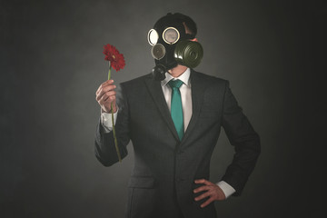 Man in gas mask and suit is holding in hands a red gerbera flower on a gray background. Pollution...