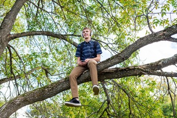 Happy student sitting on a tree branch while listening to music through his headphones.