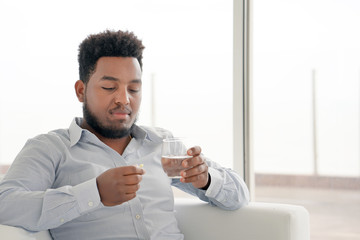 African man holding pill glass of water at home on the sofa. Sick depressed unhealthy man, about to take antidepressant pill, emergency contraceptive, painkiller for painful periods.