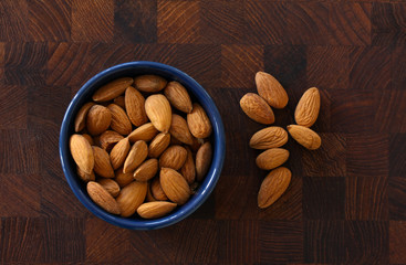 Almonds lying in a blue bowl on dark wooden background with almonds scattered around. Top view image