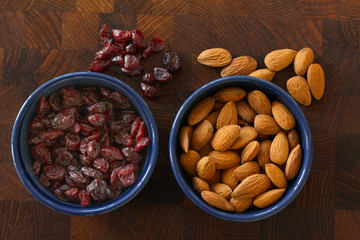 Almonds and cranberries lying in  blue bowls on dark wooden background with almonds and cranberries scattered around. Top view image.