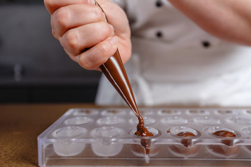 Chocolatier pours chocolate into molds. Chef in white apron using pastry bag filling hot melt...