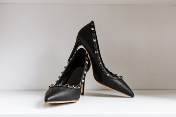 pair of black high-heeled shoes with pointed toes, decorated with metal inserts against a shelf in...