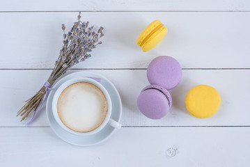 Fototapeta na wymiar Sweet delicious violet and yellow macarons and cup of latte or americano and branch of fragrant lavender on white wooden background.