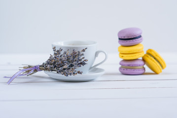 Delicious violet and yellow macarons and cup of latte or americano and branch of fragrant lavender on white wooden background.