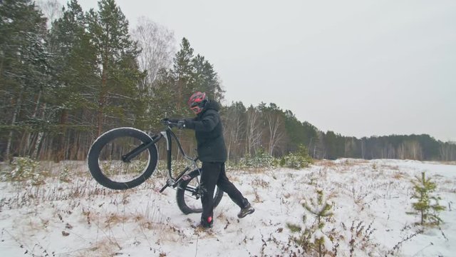 Professional extreme sportsman biker lift a fat bike in outdoor. Cyclist keep in the winter snow forest. Man walk with mountain bicycle with big tire in helmet and glasses. Slow motion in 180fps.