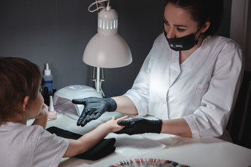 Manicurist in a black mask and black gloves looking at the nails of a little girl. Nail care of the...