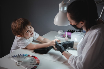 A little girl in a beauty salon. Looks at the manicurist in a black mask and black gloves. Nail...