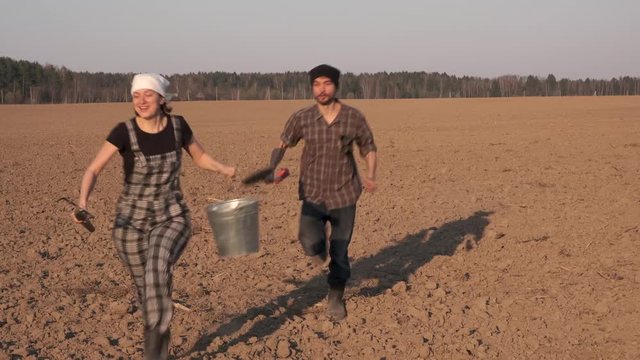 Two funny farmers man and woman running on freshly plowed field. Carry a shovel, pitchfork and bucket. Sowing season concept.