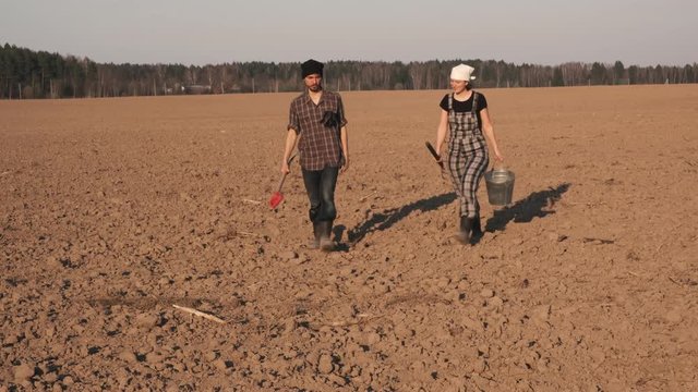 Two farmers man and woman with shovel, pitchfork and bucket walks through the freshly plowed   field and talks to each other, moving to the camera.