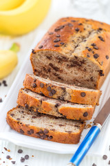 Delicious homemade banana bread with chocolate chips sliced on a white serving tray with ingredients at the background