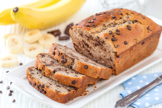 Delicious homemade banana bread with chocolate chips sliced on a white serving tray with ingredients at the background