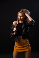 Young beautiful woman wearing in a black leather jacket, black top, short skirt and boots  posing over black background. Studio shot of  blonde woman against black background