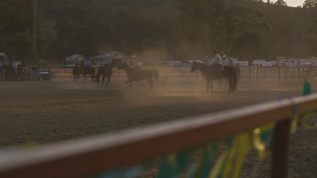 A still wide shot of people riding their horses on a small field surrounded by metal fences with the dust and dirt filling the air