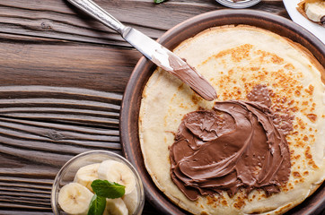 Flat lay French crepes with chocolate sauce and banana in clay bowl on wooden kitchen table