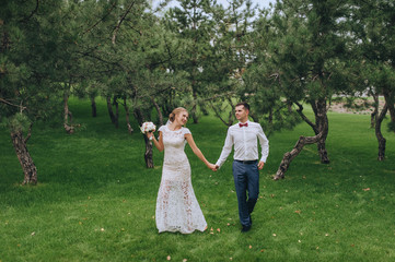 Naklejka na ściany i meble Stylish groom in shirt and beautiful blonde bride are walking, holding hands, in the forest, against the background of green grass and trees. Wedding portrait of smiling and cheerful newlyweds.